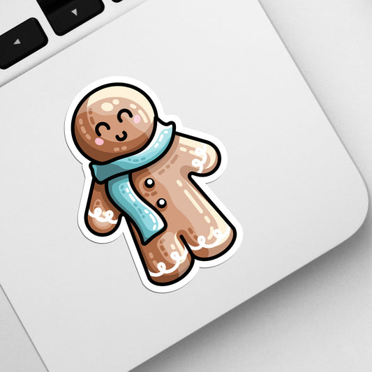The corner of a laptop keyboard with a die cut vinyl sticker of a cute gingerbread person wearing a blue scarf stuck onto it