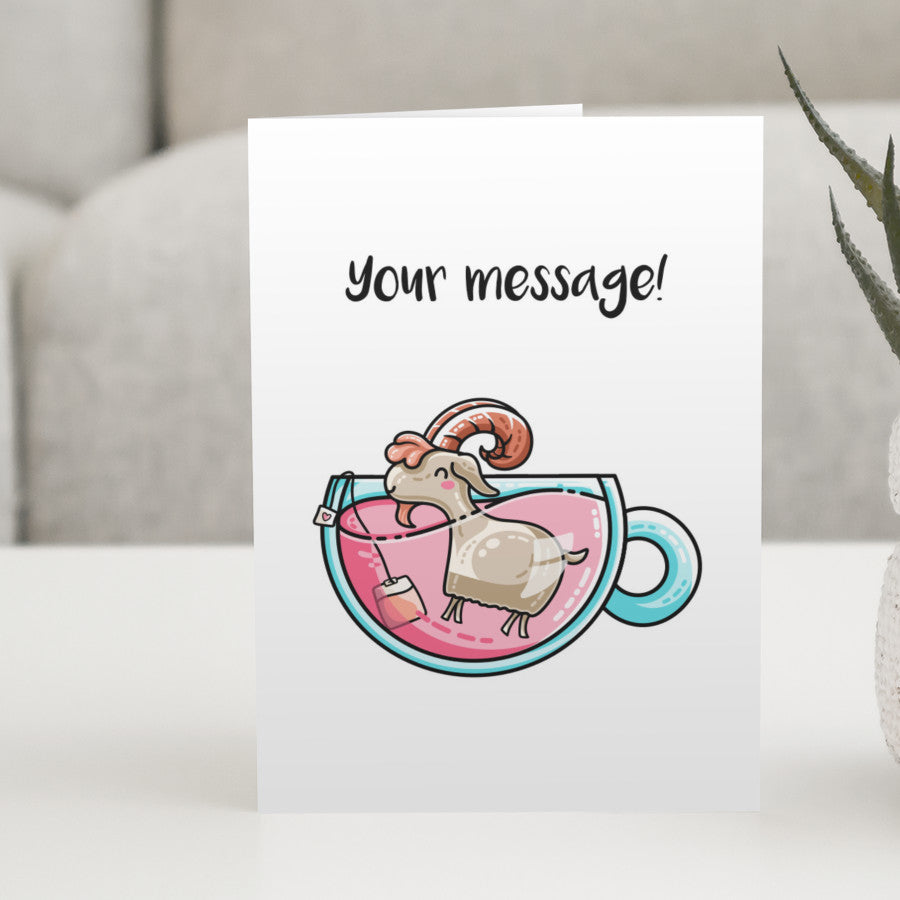 A white greeting card standing on a white table, with a design of a cute goat in a teacup of pink liquid with personalised wording above