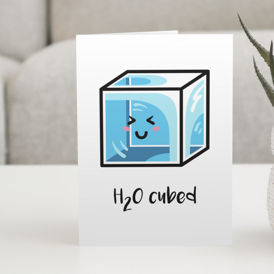 A white greeting card standing on a white table, with a design of a kawaii cute blue cube of ice and the words 'H20 cubed'