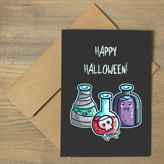 A dark grey greeting card with the words happy halloween, lying flat on a brown envelope, with a design of three glass bottles in halloween costume as a mummy wrapped in bandages with green liquid, a vampire in a black cape with purple liquid, and a skull with bones in red liquid