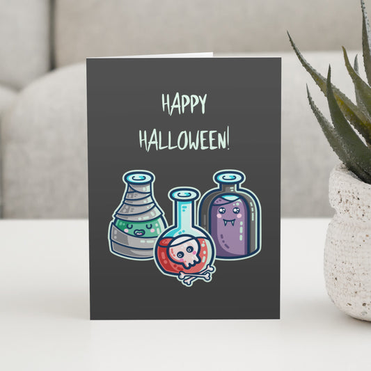 A dark grey greeting card with the words happy halloween, standing on a white table, with a design of three glass bottles in halloween costume as a mummy wrapped in bandages with green liquid, a vampire in a black cape with purple liquid, and a skull with bones in red liquid