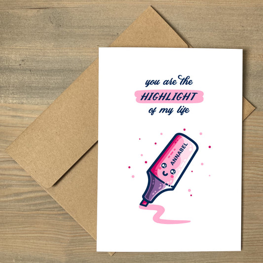 A white card lying flat on a brown envelope. Design on the card is of a pink kawaii cute highlighter pen with a smiling face and the name 'Emma' written on it and the words 'you are the highlight of my life' written above. The word 'highlight' is highlighted in pink.