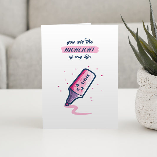 A white card standing on a table. Design on the card is of a pink kawaii cute highlighter pen with a smiling face and the name 'Emma' written on it and the words 'you are the highlight of my life' written above. The word 'highlight' is highlighted in pink.