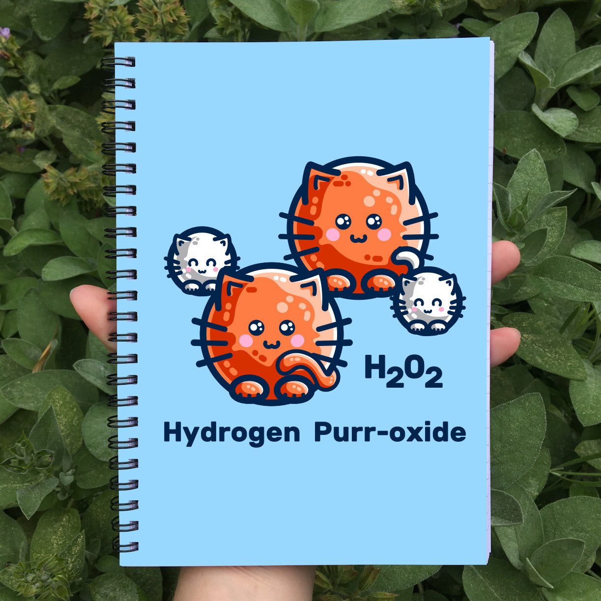A blue spiral notebook being held in a hand with a design on the front cover of four cats representing the atoms in a hydrgen peroxide molecule with the words H202 hydrogen purr-oxide