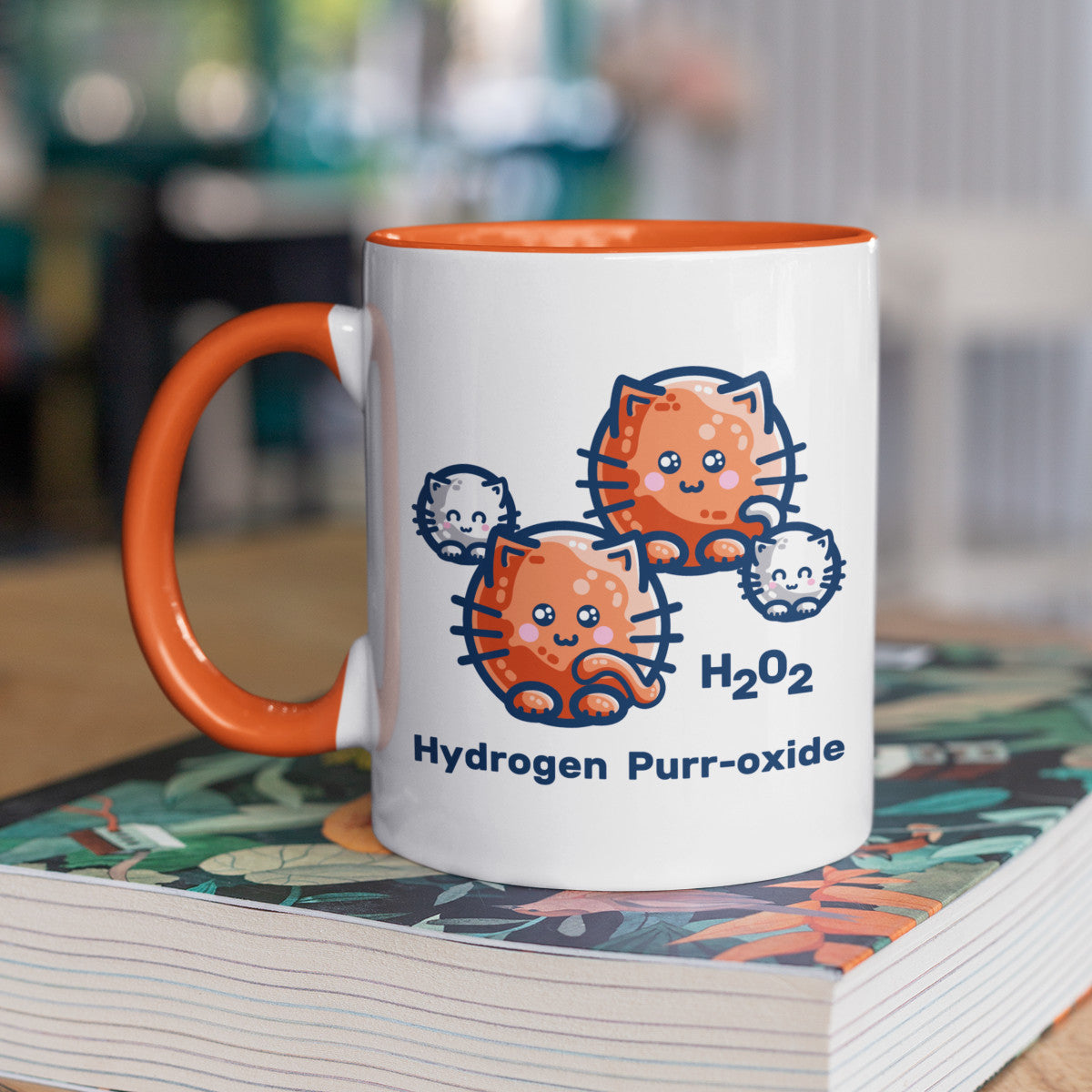 A two toned white and orange ceramic mug standing on a book and with the handle to the right, showing a design of a hydrogen molecule with the hydrogen atoms replaced by round white kittens and the oxygen atoms replaced by larger round ginger cats and the words H202 hydrogen purr-oxide.