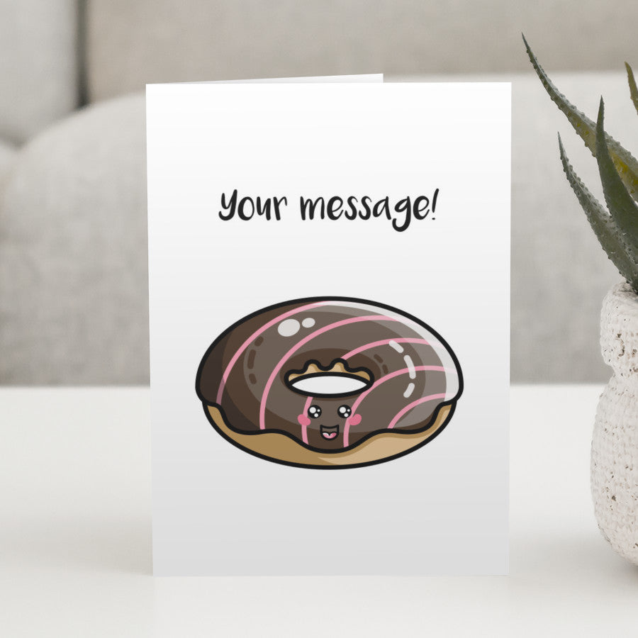 A white greeting card standing on a white table, with a design of a kawaii cute chocolate iced doughnut with your own personalised message above.