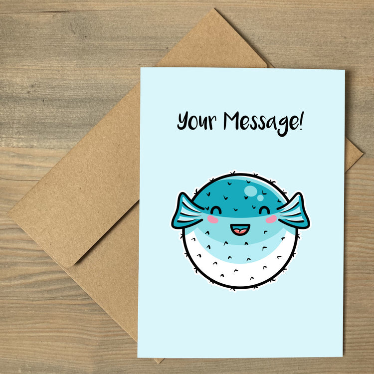A pale turquoise greeting card lying flat on a brown envelope, with a design of a kawaii cute turquoise and white puffer fish with personalised wording above