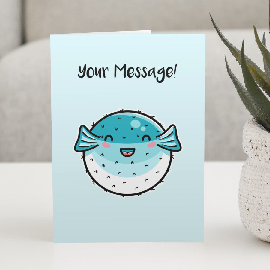 A pale turquoise greeting card standing on a white table, with a design of a kawaii cute turquoise and white puffer fish with personalised wording above