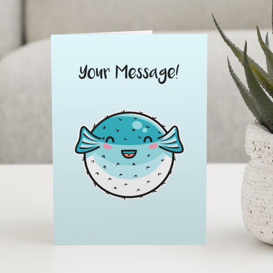 A pale turquoise greeting card standing on a white table, with a design of a kawaii cute turquoise and white puffer fish with personalised wording above