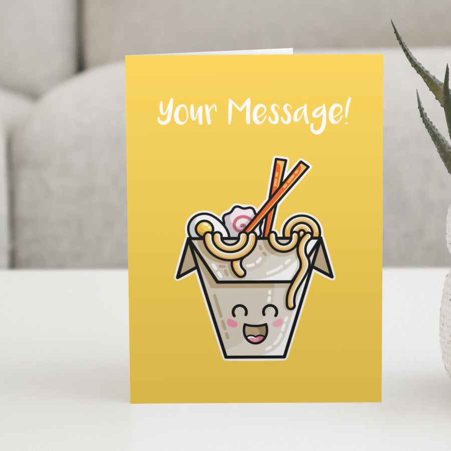 A yellow greeting card standing on a white table, with a design of a kawaii cute takeaway box of ramen noodles and chopsticks with a personalised message above