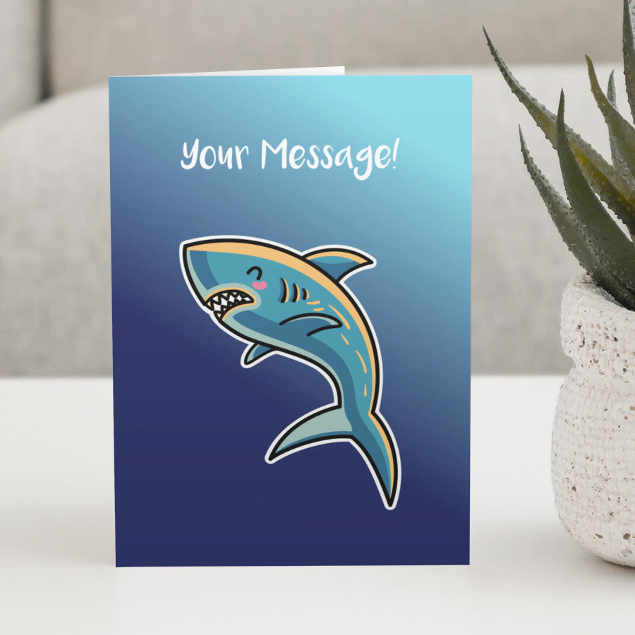 A blue greeting card standing on a white table, with a design of a kawaii cute shark with personalised wording above