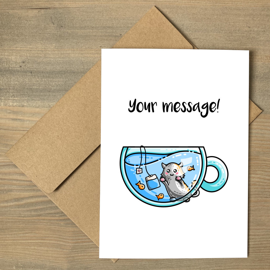 A white greeting card lying flat on a brown envelope, with a design of a glass teacup of blue liquid with orange fish and a tea bag in it, with a cat on the far side gazing in at them and a personalised message above.