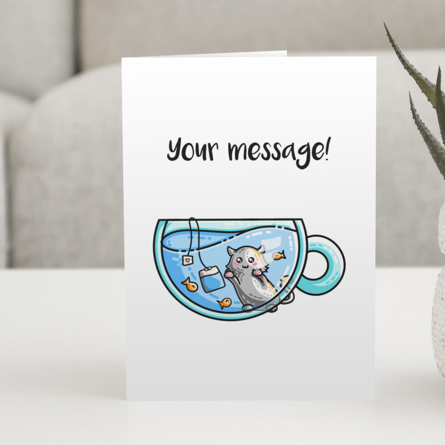 A white greeting card standing on a white table, with a design of a glass teacup of blue liquid with orange fish and a tea bag in it, with a cat on the far side gazing in at them and a personalised message above.