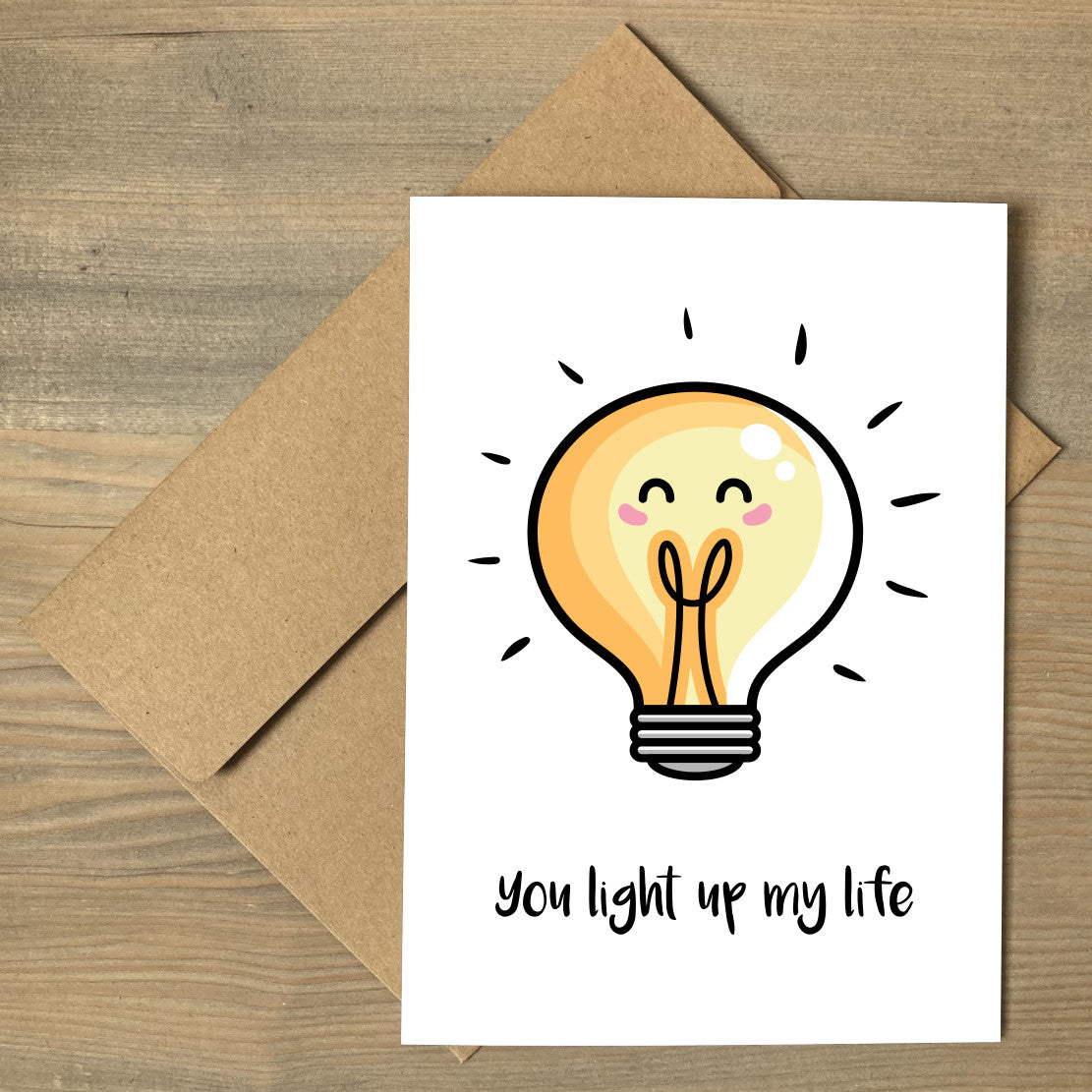 A white greeting card lying flat on a brown envelope, with a design of a kawaii cute lightbulb with words beneath reading 'you light up my life'