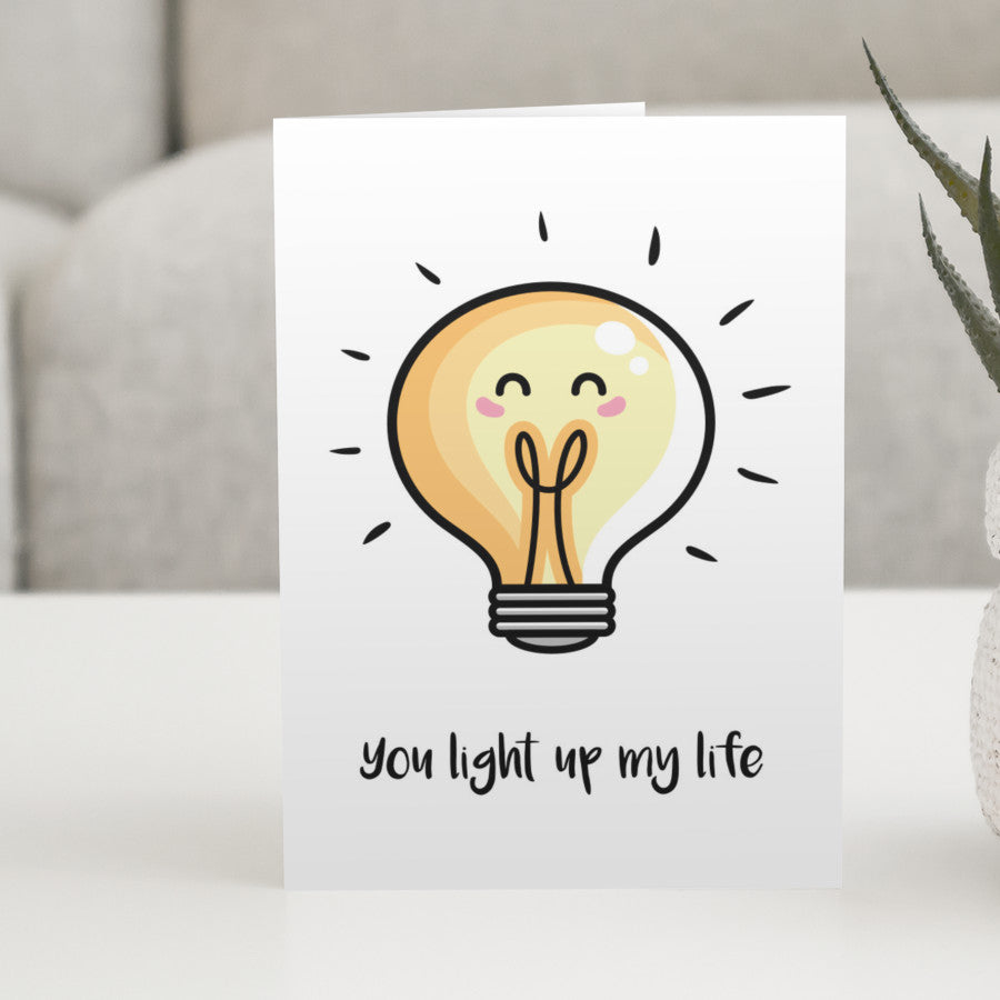 A white greeting card standing on a white table, with a design of a kawaii cute lightbulb with words beneath reading 'you light up my life'