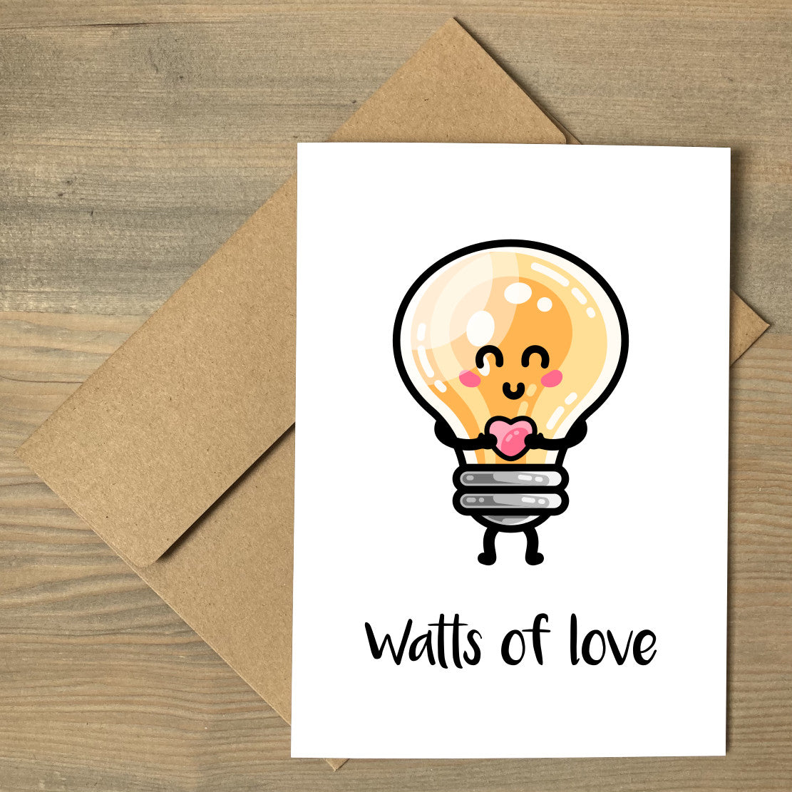 A white greeting card lying flat on a brown envelope, with a design of a kawaii cute lightbulb holding a heart and the words watts of love beneath