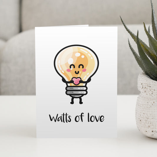 A white greeting card standing on a white table, with a design of a kawaii cute lightbulb holding a heart and the words watts of love beneath