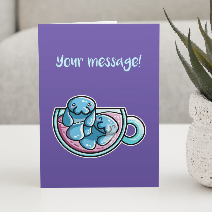 A purple greeting card standing on a white table, with a design of two kawaii cute blue manatee swimming in a teacup, with a personalised message above
