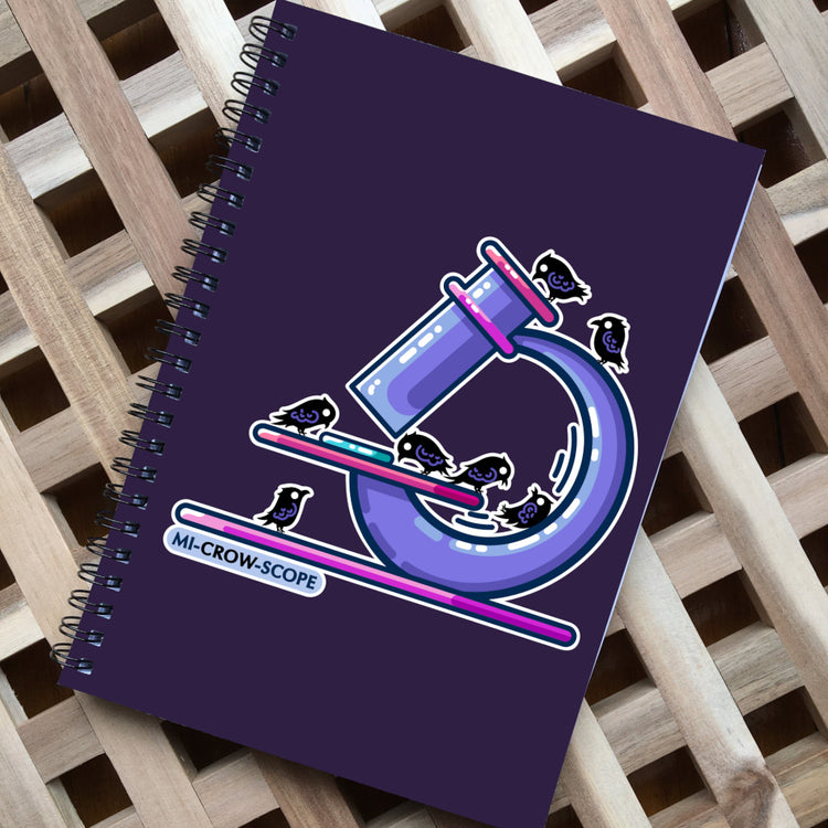 A dark purple spiral bound notebook, with black spiral wire. Design on the front of a pink and purple microscope with 7 crows standing on it and the pun word mi-crow-scope written beneath in capital letters.
