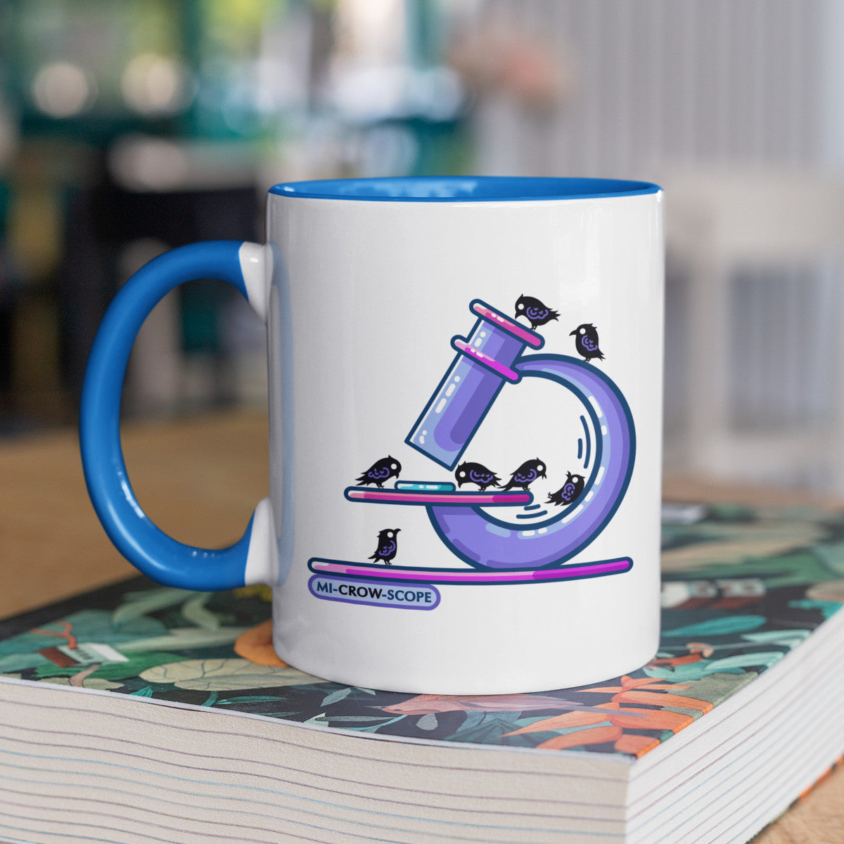 A two-toned white and blue ceramic mug standing on a book with a design printed on it of a purple microscope with crows standing on it.