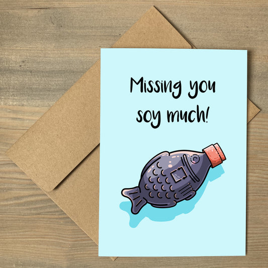 A pale blue greeting card lying flay on a brown envelope with a design of a kawaii cute soy with red lid facing diagonally to the top right and full of dark soy sauce with the words missing you soy much written above