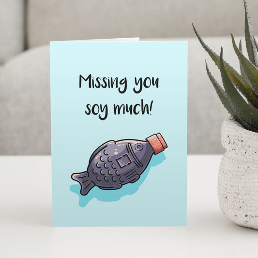 A pale blue greeting card standing on a white table with a design of a kawaii cute soy with red lid facing diagonally to the top right and full of dark soy sauce with the words missing you soy much written above