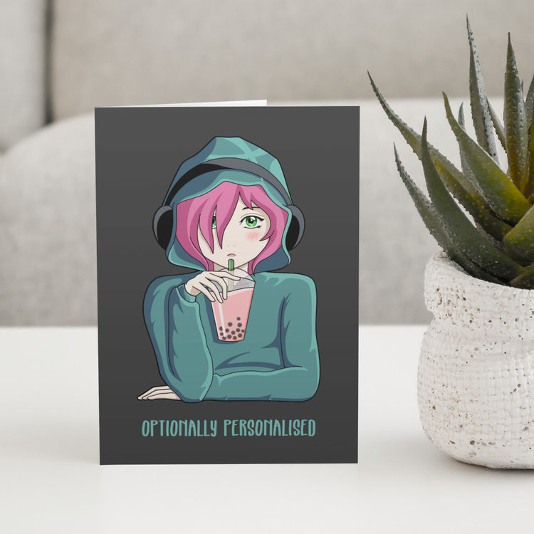 A dark grey greeting card standing on a white table with a design of a pink haired anime girl wearing a green hoodie and black headphones drinking pink boba bubble tea