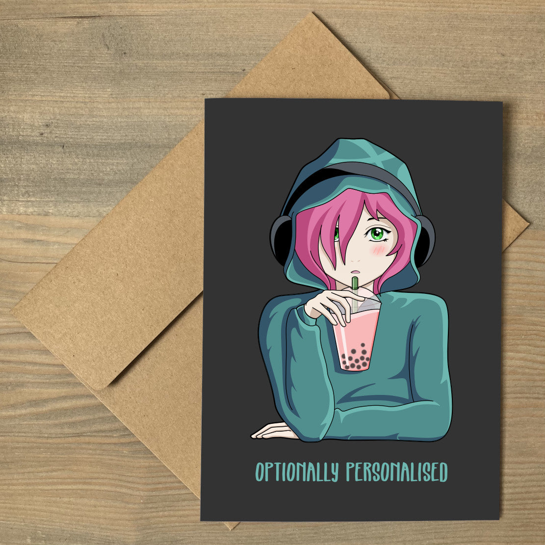 A brown envelope beneath a dark grey greeting card with a design of a pink haired anime girl wearing a green hoodie and black headphones drinking pink boba bubble tea