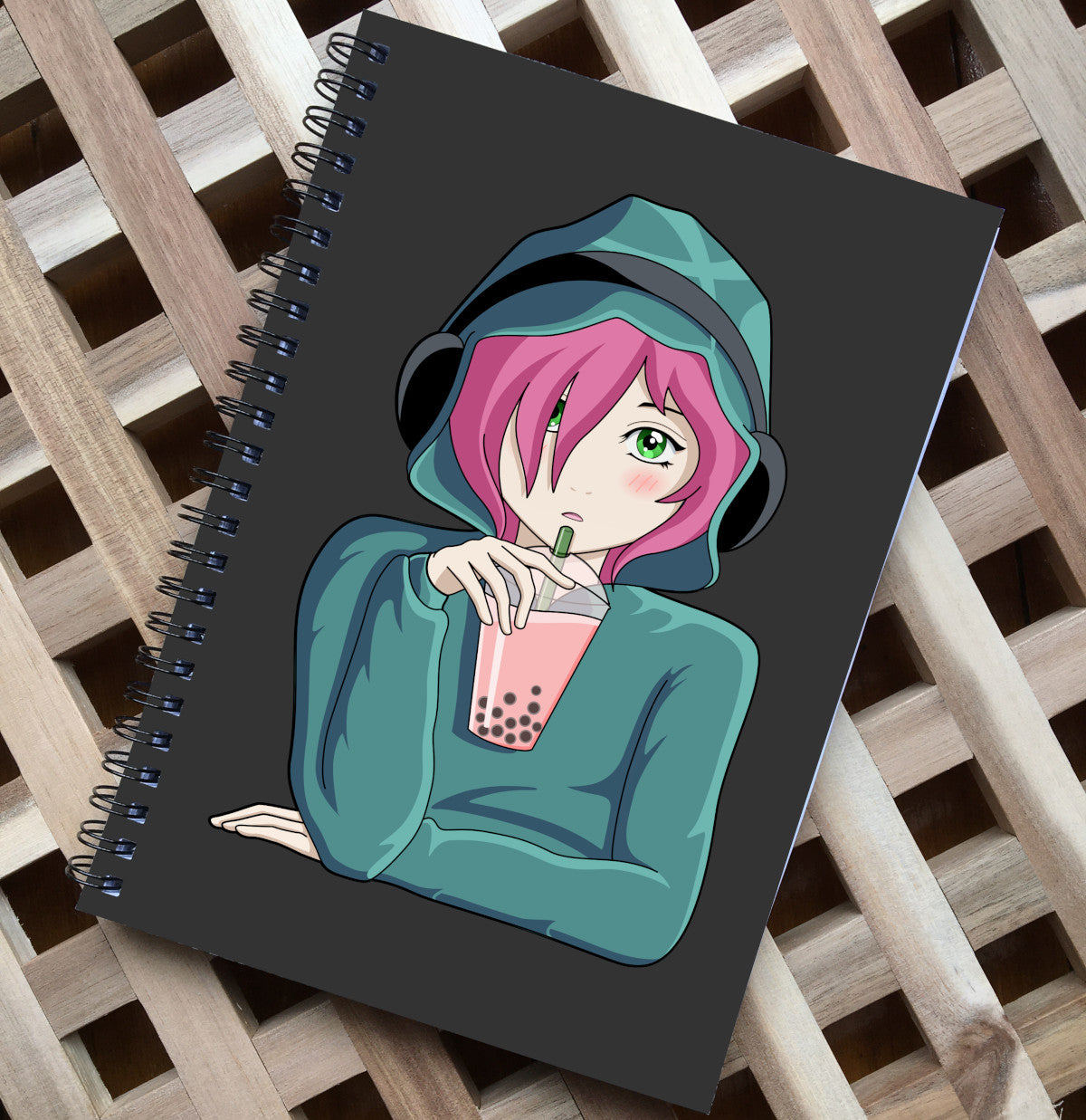 Spiral notebook lying flat on a wooden surface, has a dark grey front over featuring an anime girl wearing a green hoodie with the hood up and black headphones over the top, she has green eyes and pink hair and is holding a transparent plastic cup of pink boba with a green straw in her right hand which has her elbow resting on a surface, her left arm and hand is lying flat across her body in front of her on the surface as if she is sitting at a table
