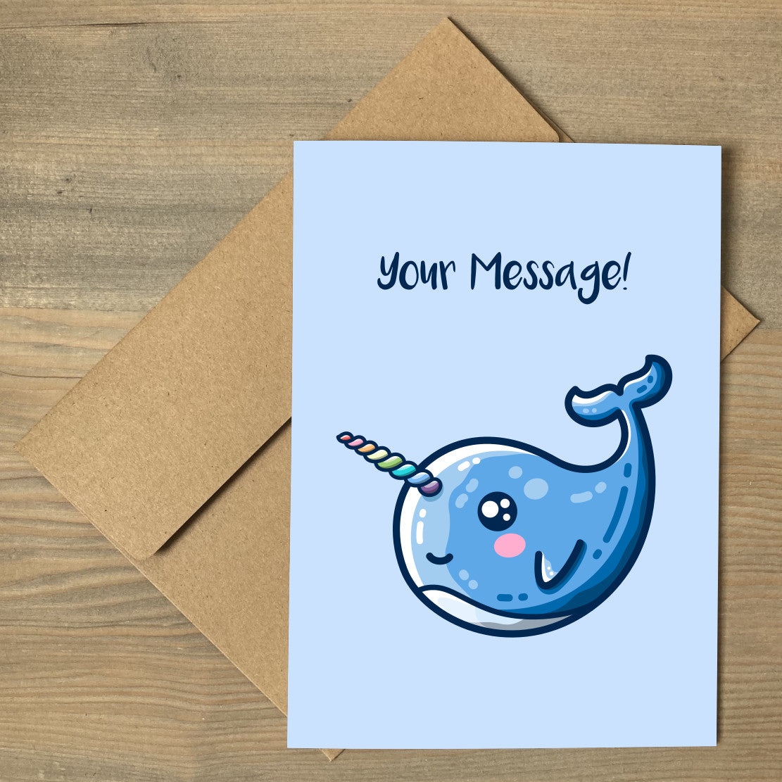 A brown envelope beneath a pale blue greeting card with a design of a kawaii cute blue narwhal facing to the left and with a rainbow striped horn and the words your message above