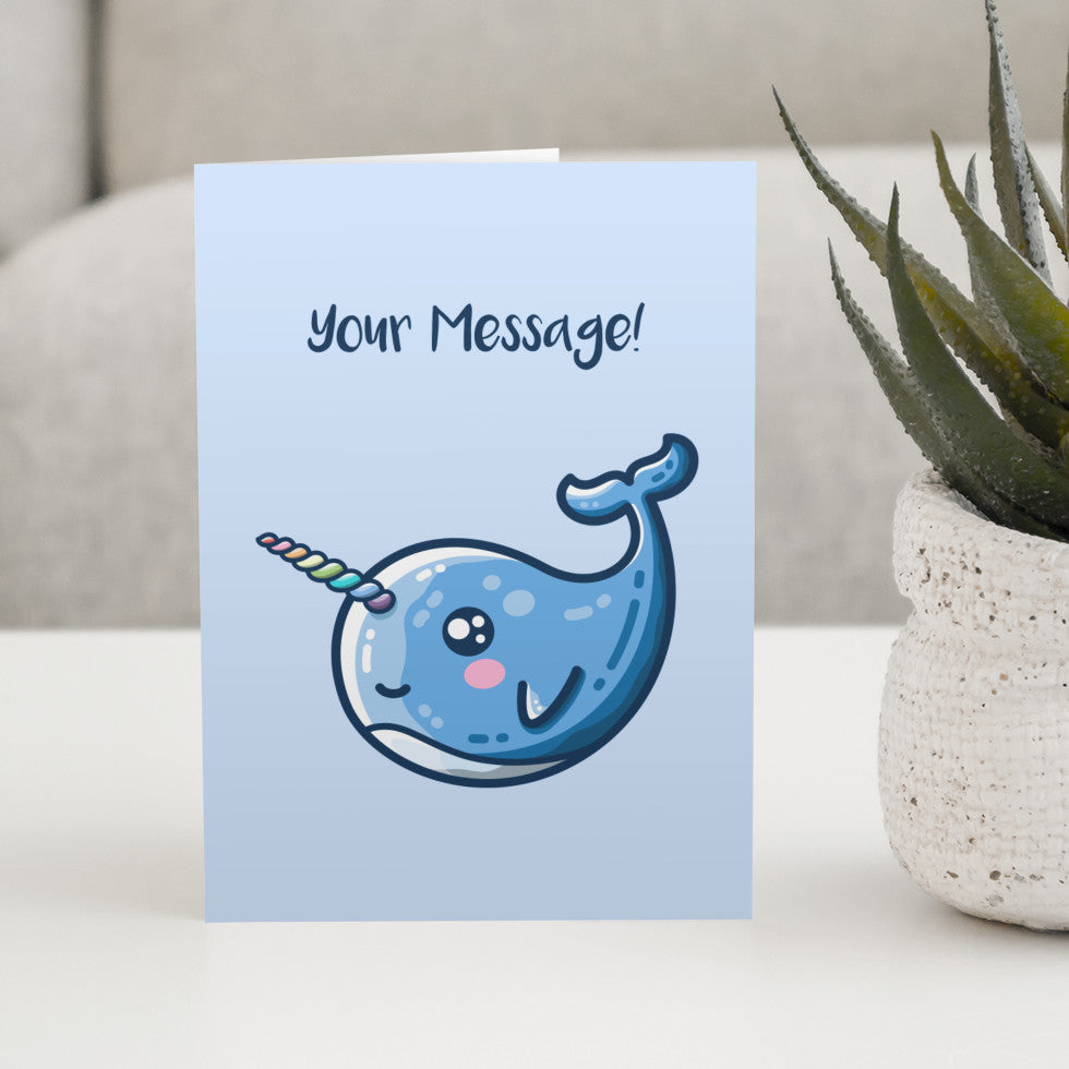 A pale blue greeting card standing on a white table with a design of a kawaii cute blue narwhal facing to the left and with a rainbow striped horn and the words your message written above