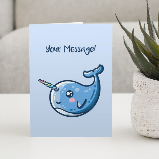 A pale blue greeting card standing on a white table with a design of a kawaii cute blue narwhal facing to the left and with a rainbow striped horn and the words your message written above
