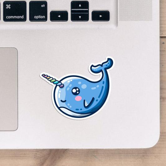 A shaped vinyl sticker of a smiling kawaii cute blue narwhal with a twisted rainbow striped horn, shown stuck onto the bottom right hand corner of a laptop computer keyboard