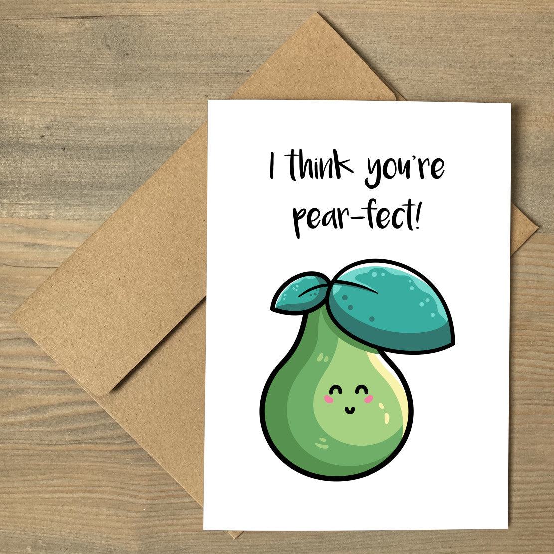 A white greeting card lying flat on a brown envelope, with a design of a kawaii cute green pear and two leaves with wording above reading I think you're pear-fect!