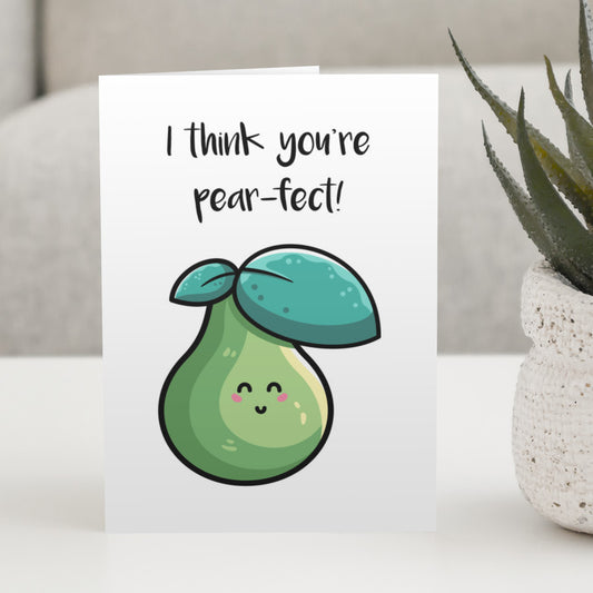 A white greeting card standing on a white table, with a design of a kawaii cute green pear and two leaves with wording above reading I think you're pear-fect!