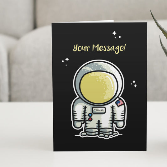 A black greeting card standing on a white table with a design of a cute astronaut space suit with a personalised name on its chest and a personalised message above.