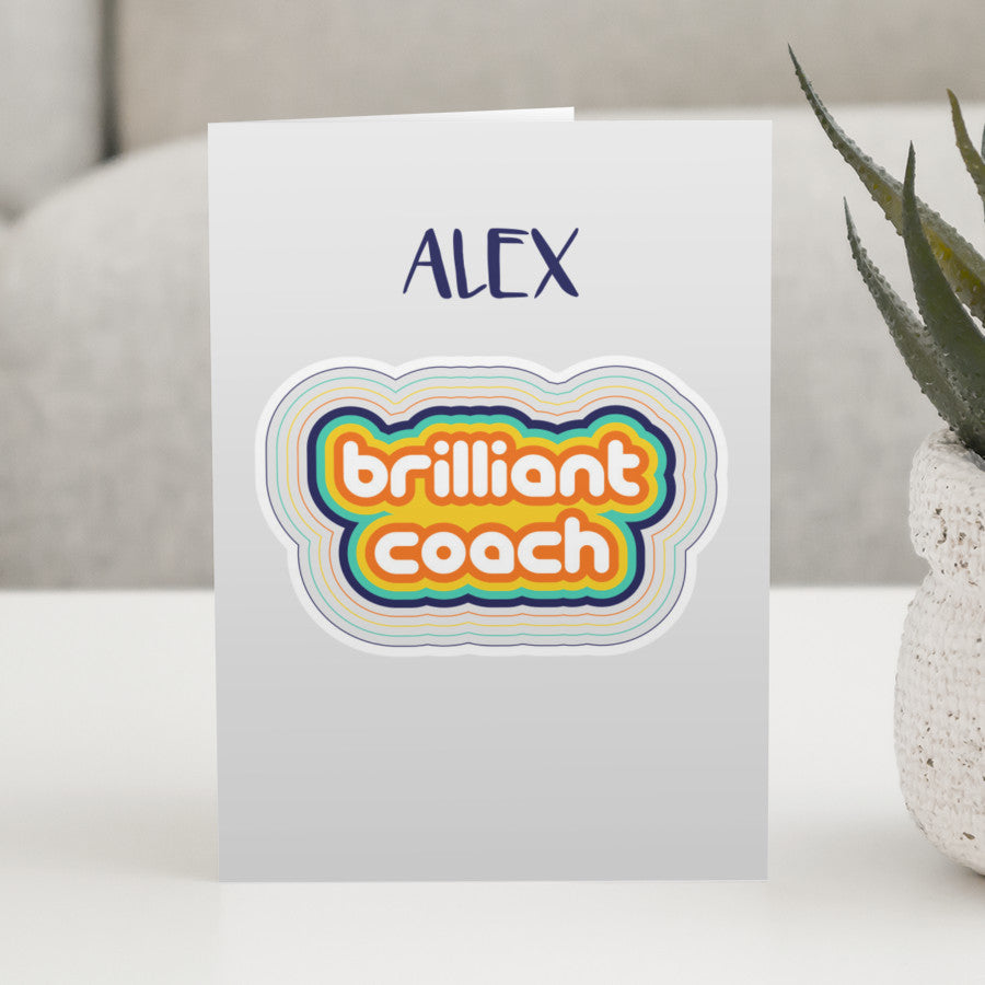 A grey greeting card standing on a white table with a design of colourful stripes surrounding the words brilliant coach, personalised with a name above.