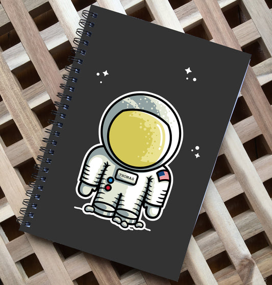 Black spiral notebook lying flat on a wooden surface, has a cute astronaut design printed on the center of the front cover with the name Thomas on its chest plate and some clusters of small white stars above and around the helmet