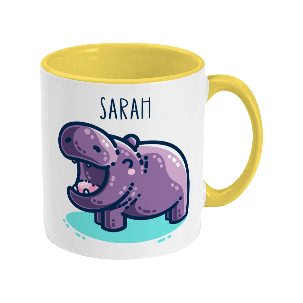 A white ceramic mug with a yellow handle on the right and yellow inside. A name is written in thin dark blue upper case letters above a kawaii cute purple hippo with a thick dark blue outline and a turquoise shadow beneath. The hippo is seen side on facing to the left and looks happy with its mouth open wide.