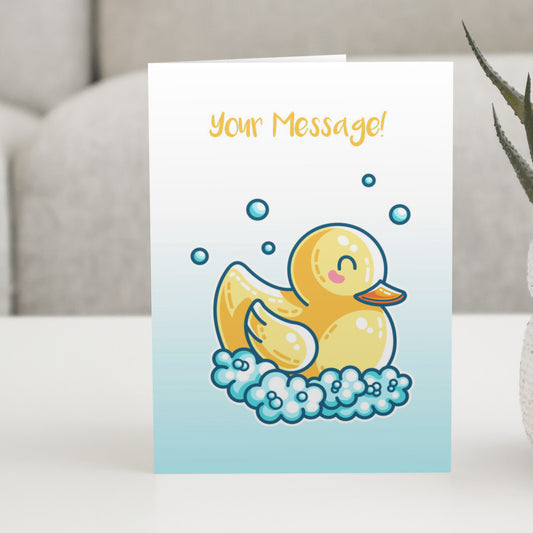 A white to blue gradiant greeting card standing on a white table, with a design of a kawaii cute yellow rubber duck surrounded by bubbles and personalised with your message above