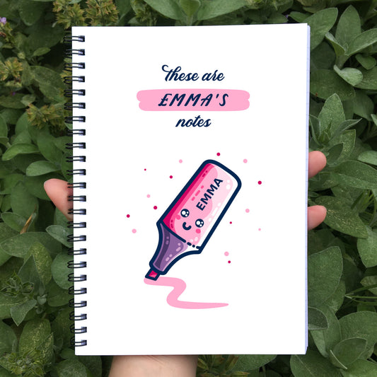A white spiral bound notebook held in a hand showing the front cover. On the front cover are the words 'these are Emma's notes' above a drawing of a kawaii cute happy pink highlighter pen at an angle drawing a thick pink line and also personalised with the name Emma on the pen.