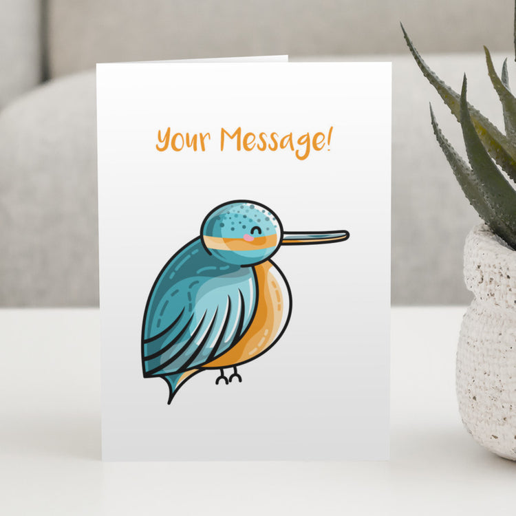 A white greeting card standing on a white table, with a design of a cute turquoise and orange kingfisher and a personalised message above