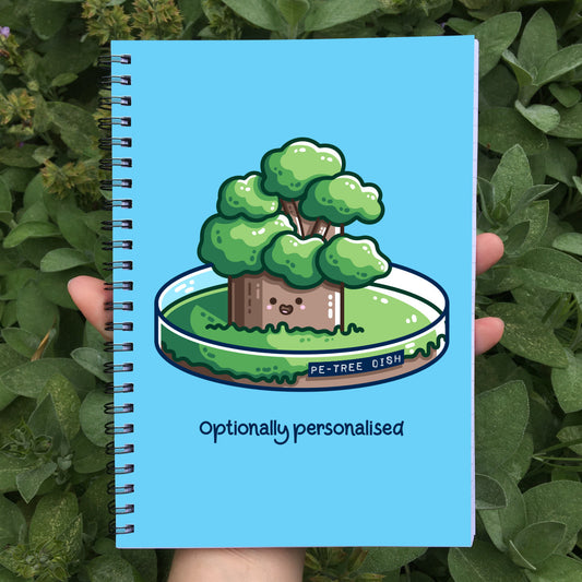 A blue spiral notebook held in a hand, with a picture on the front of a kawaii cute tree growing in a petri dish and beneath it the words 'optionally personalised'