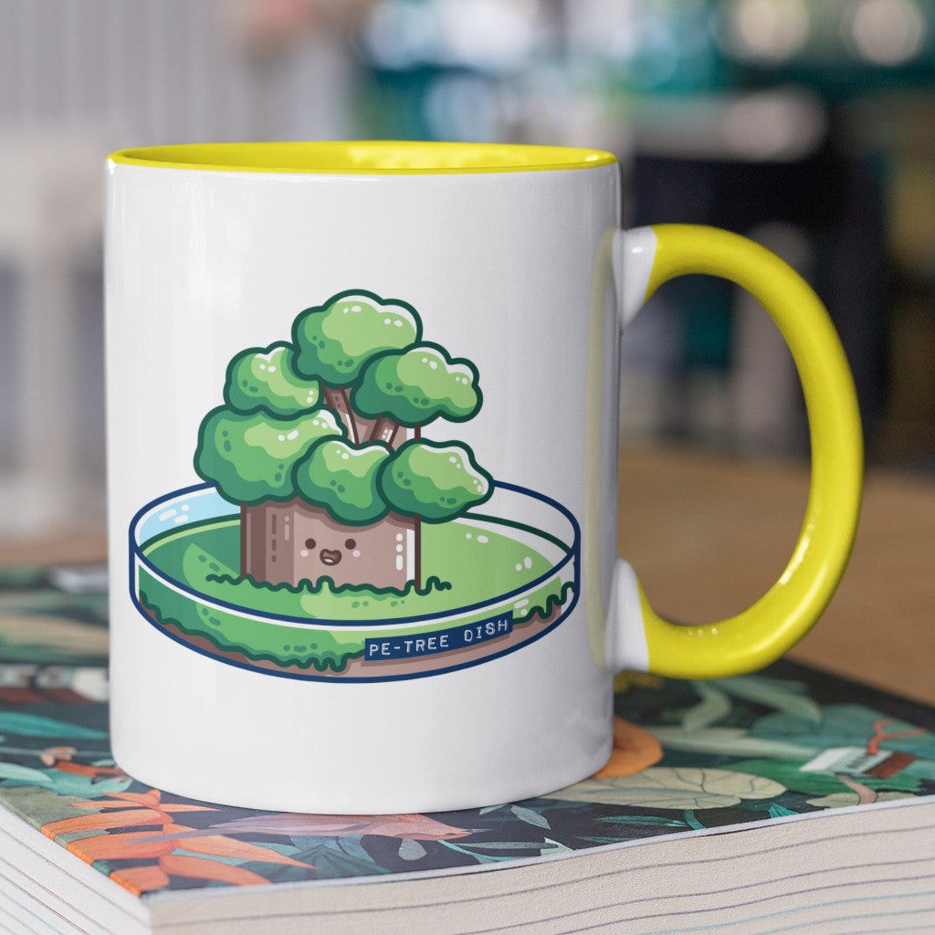 A two toned white and yellow ceramic mug with the handle to the right and standing on a book, showing a design of a kawaii cute tree growing in a petri dish.