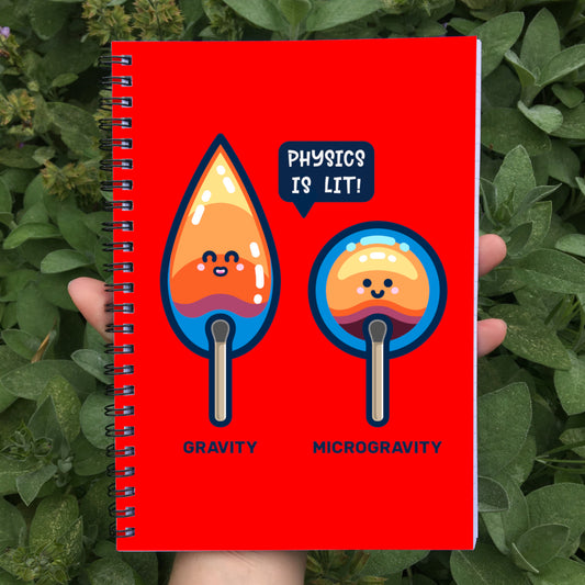 Held in a hand is a closed spiral notebook showing red front cover with a design of 2 cute flames, one pointed with word gravity and one round with word microgravity and speech bubble saying physics is lit!