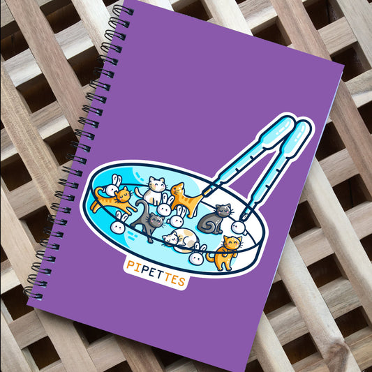 A purple spiral notebook, with black spiral wire, on a slatted wooden surface. The front cover has a picture of a petri dish containing cats and rabbits and two pipettes, with the word pipettes written beneath.