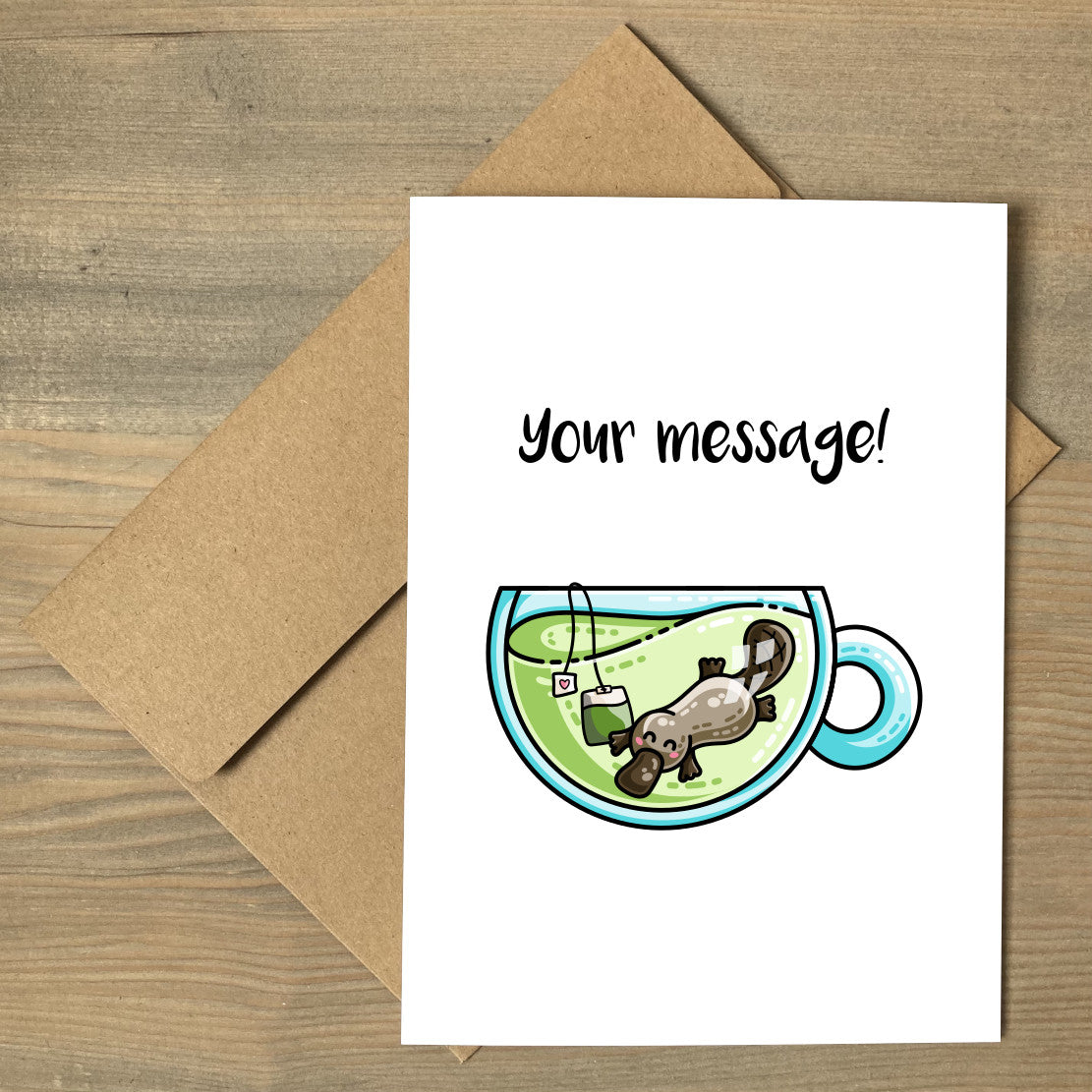 A white greeting card lying flat on a brown envelope, with a design of a cute platypus swimming in a glass teacup of green tea with personalised wording above