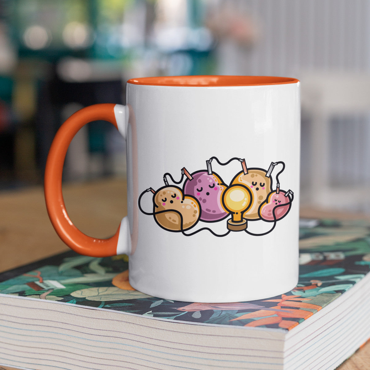 A two-toned white and orange ceramic mug, standing on a book, with a design of a potato battery of 4 potatoes asleep around a light bulb they are powering