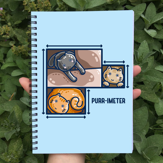 A pale blue spiral notebook held in a hand showing the front cover which features three cute cats in adjoining cardboard boxes seen from directly above, with measurement lines around the edges and the word 'purr-imeter'.