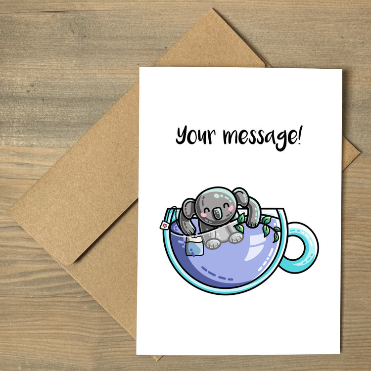 A white greeting card lying flat on a brown envelope, with a design of a glass teacup of lilac liquid with a koala bear and tea bag in it, and a personalised message above